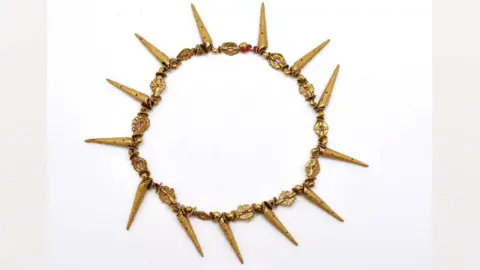 Museum of Archaeology and Anthropology A 19th Century necklace from Asante, Ghana. It is intricately worked in rich yellow gold with flat and round gold beads strung along in a circle, and 13 long gold spikes shaped like spiral shells spacing out the beads every seventh bead