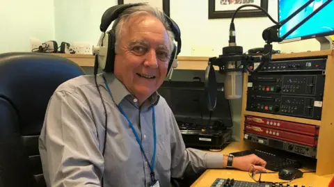 John Alborough sits in a radio studio with headphones on. He is looking at the camera and smiling. He has grey hair and wears a blue shirt. In front of his face is a radio microphone and other radio equipment.