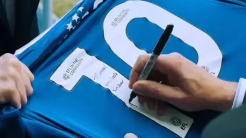 Reform UK A still from a video showing a hand signing the back of a blue football shirt that has the number 10 on it.