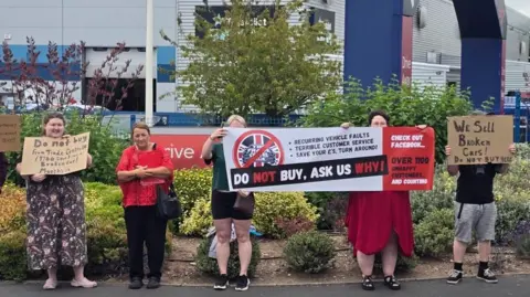 Soo Townsend Seven people, a mixture of men and women, stand on the pavement outside a large building belonging to Trade Centre UK. They are holding signs which include the words "do not buy, ask us why!"