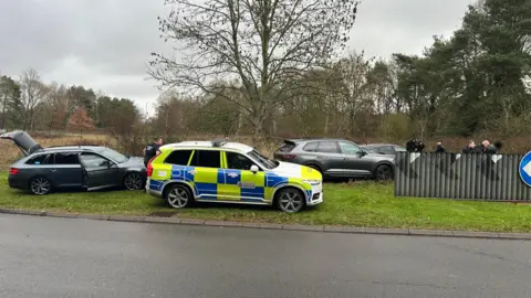 A yellow and blue police vehicle sits on a grassy bank beside the A46 in Warwickshire. Three other black vehicles are parked nearby. Police officers can be seen behind a road barrier. 