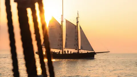 The Sprit of Falmouth sailing at sea in the sunset. It is a large ship with three sails and you can see the silhouettes of people aboard. 