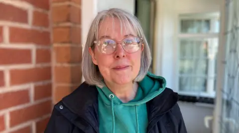 A woman with shoulder-length grey hair, wearing a green hoodie and black jacket, stands on a doorstep that leads into a house porch.