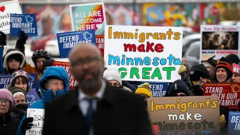 Getty Images Warmly dressed demonstrators rally outside a shop. They are holding signs in praise of migration including "Immigrants make Minnesota great".
