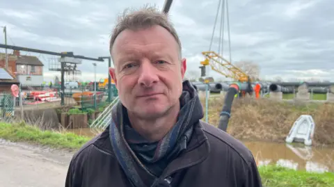 Ian Withers. He has short brown hair and no facial hair. He is pictured outside, standing in front of a station to pump flood water away.