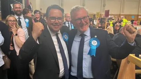 Craig Hoy and David Mundell with blue rosettes punch the air in celebration at the election count in Dumfries