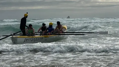 Five people in a boat with long oars riding across the surf