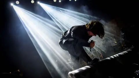 A man crouches on stage. He is holding a microphone and wearing all black. He has long, dark hair. His upper-body is parallel to the ground, and he is illuminated by two spotlights from above. 