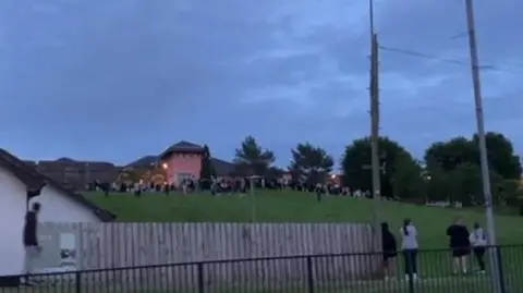 A number of young people can be seen congregating on a grassy embankment near a large pink building. Telephone wires and a picket fence can be seen at the bottom of the hill close to a white building.
