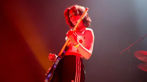 A woman in a white top and black tracksuit bottoms playing guitar