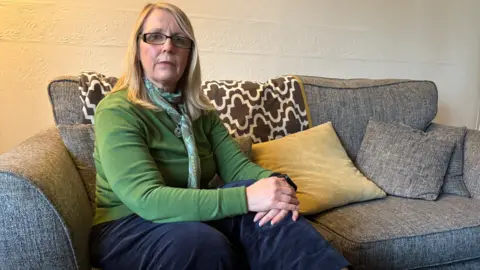 A woman wearing a green top and jeans sits on grey sofa. Her hands are clasped together and lying across her left knee. A number of cushions can be seen on the sofa behind her.