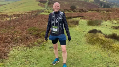 Matt Sears A man with a shaved head stands on an expanse of grass and heathland. He is wearing trainers, black running shorts, a black running top and black gloves and has a rucksack on his back. He has a sign pinned to his front with the number 180 on it.