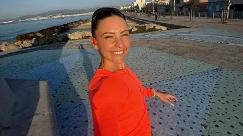 Alysia Mcandrew Alysia stands on a Promenade, possibly abroad and smiles at the camera. She is wearing an orange running top with long sleeves and has a pair of sunglasses balanced on her head as she smiles at the camera.