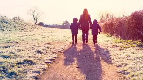 Getty Images A woman with her back to the camera walks down a field path, holding the hands of two children. They are all wearing trousers and dark winter coats and there is frost on the ground.