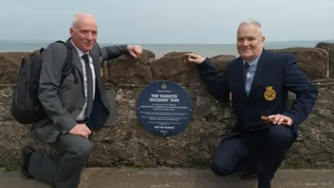 Andrew Bannister Two men in suits kneeling down next to a blue circle plaque on the wall on the coast line. The plaque is remembering the 1949 Yangtze Incident.
