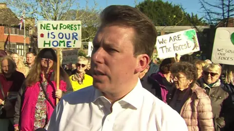 James Hartley-Binns stood in a suit in front of crowds of people protesting and holding signs.