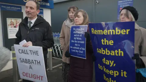 A man and three women protest  holding placards outside Tesco. One reads: "Call the Sabbath a delight".