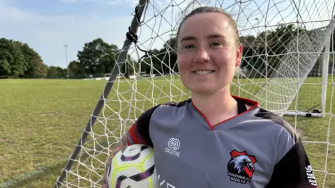 A lady stood with a ball under her arm in front of a football net with a Ravenshead Red shirt on.