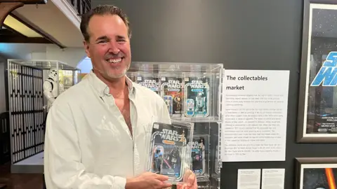 Matt Fox wears a white shirt and is standing in front of a wall of boxed star wars figurines. He is holding a Darth Vader box.  