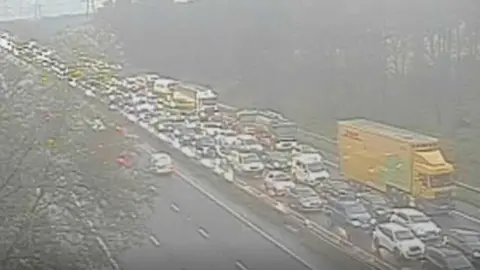 A large queue of cars and a yellow lorry on the M56. The conditions are wet, with poor visibility. The opposite carriageway is clear.