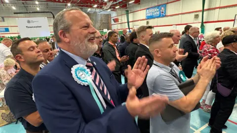 Martin Heath/BBC James Petter with short dark hair and grey beard, wearing a blue jacket, light blue shirt and pink and black tie, with a light blue reform rosette. He is smiling and clapping alongside other people similarly occupied at an election count.