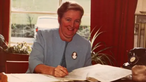 Family Handout Margaret Grey around middle age when she was a headmistress. She is wearing a blue blazer with a female silhouette brooch on her left lapel. She is sitting behind a wooden desk writing on a stack of papers. To her left is a black rotary telephone and behind her is a big window with lots of plants on the windowsill. She has her blonde hair tied back and is smiling at the camera.