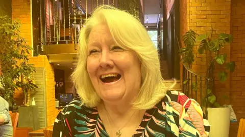 Lilian Weimer Lilian Weimer with shoulder-length light-coloured hair sitting at a table in a restaurant or café, hands clasped, wearing a patterned top and jewellery, with indoor plants, brick walls, and a staircase visible in the background.