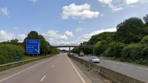 Junction four of the M54, a single red car is visible in the distance on the eastbound carriageway on the left, while cars and a lorry approach on the westbound carriageway on the right. A footbridge crosses the lanes, with trees bordering the motorway