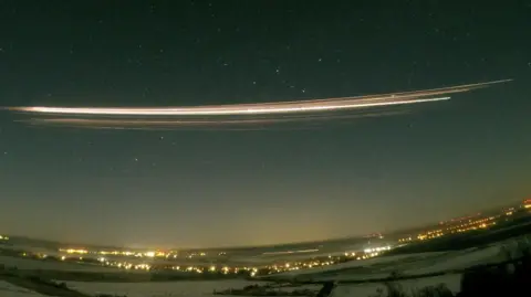 A photograph using 30-second exposure showing the Falcon 9 upper stage re-entering the atmosphere above Berlin, Germany. A long streak of bright yellow and white is seen in the dark night sky, above a landscape of dark field and bright lights from the city.