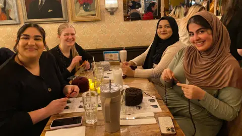 A group of four women are sitting around a table in a restaurant. They all have drinks on the table along with their phones as they learn to crochet with yarn