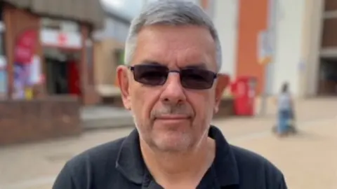 BBC Pete is looking at the camera with dark glasses on and a black T'shirt.