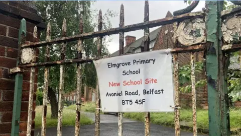 Rusted green and white railings have a laminated piece of paper attached. The piece of paper says 'Elmgrove Primary School, New School Site, Avoniel Road, Belfast BT5 4SF