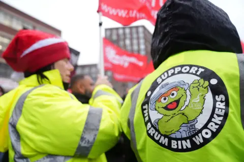 PA Media The backs of two workers wearing hi-vis jackets. One of them has a logo and 'support the Brum bin workers' written on it