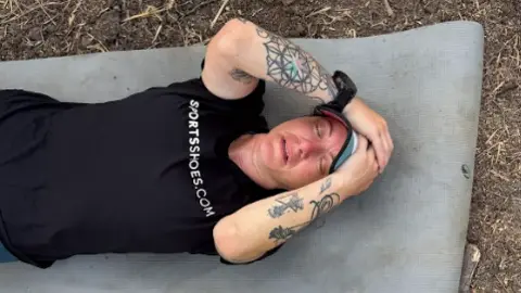 Hannah lies on yoga mat. She wears a black t-shirt and light blue sports cap and has tattoos on both arms. Her eyes are shut as though she is very tired and she is lying outside on rough ground with grass or straw.