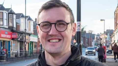 Sunderland Labour Group Michael Mordey is smiling. He is standing on a street with houses and a row of shops. He has short brown hair and brown eyes. He wears circular glasses and a black coat. 