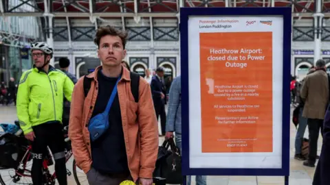 Reuters A man wearing an orange jacket, black top and blue bag looks beyond the camera to an arrivals board at Paddington station while he stands next to a white and orange sign saying 'Heathrow Airport closed due to power outage"