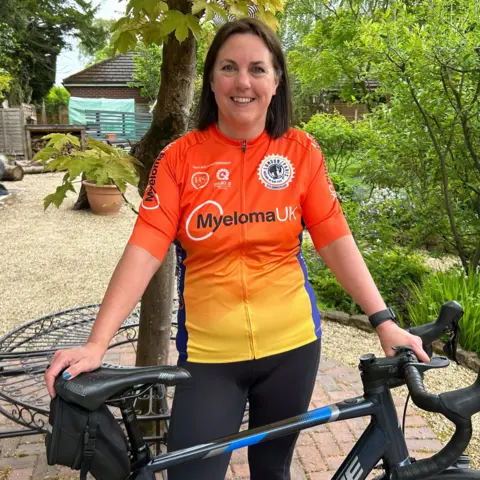 Gavin Kermack / BBC The woman from the above picturem this time wearing black lycra leggings and a bright orange and yellow lycra top with "Myeloma UK" emblazoned on the front. She is standing in a garden with her hands resting on a bicycle.