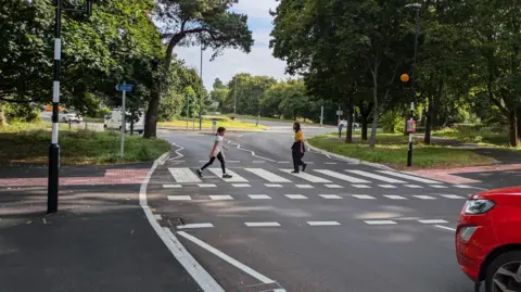 BCP The new crossing on Glenferness Avenue looking towards Talbot roundabout. Two people are seen crossing the road. Part of a red car is seen waiting. It's a sunny day.