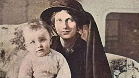 Lady Petre is dressed in all black with a hat on. A child is on her lap. She is sitting on a sofa inside.