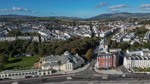 MANX SCENES An aerial view of the road along Douglas promenade, you can see the roundell in the middle and the Villa Marina on the left of the picture, there are houses in the background and hills.