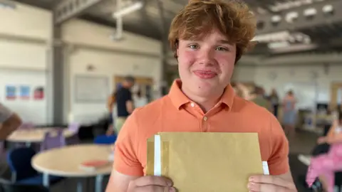 A man with orange hair smiles widely and holds an envelope 