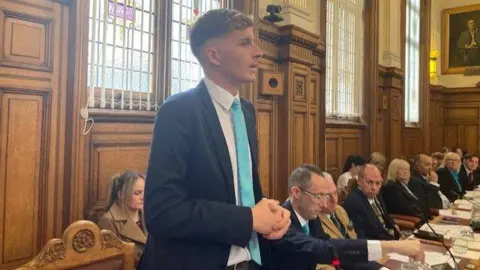 A man with short blond hair is wearing a black blazer with white shirt and light blue tie. He has a serious expression and is stood up along a long table with other people sat down at desks. It is an old building of a hall.