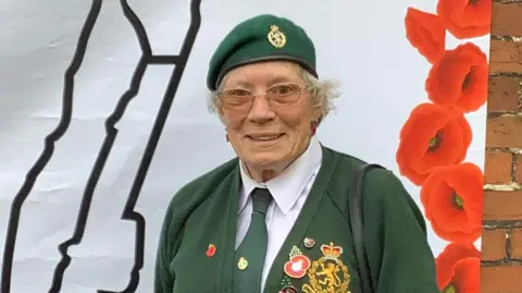 A smiling Sandy Cawley wears a green beret, tie and cardigan with army badges and insignia. She wears glasses and a white shirt, with a black handbag over one shoulder. 