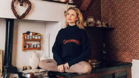 Bryony Coles Saoirse-Monica Jackson has long blonde curly hair. She is sitting on a kitchen bench and wearing a black jumper with the word Guinness written in red block letters.