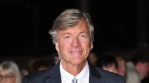 Richard Madeley has long grey hair and is smiling at the camera. He is wearing a dark suit with a white shirt.