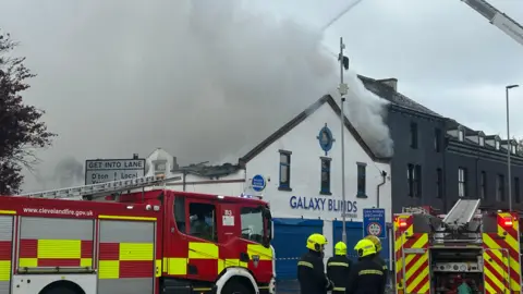 Two red and yellow fire engines parked in front of Galaxy Blinds which is a white and blue building with a pointed front. Grey smoke covered the entirety of the roof and is billowing upwards and to the right. Four firefighters are standing in front with yellow helmets on. There is a row of grey terraced buildings to the right.