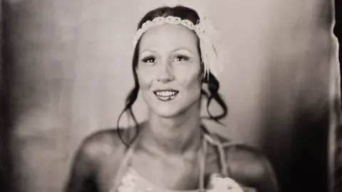 Guy Bellingham Real Amy G wearing an embroidered headband with feathers and a strappy top. She has dark hair that is tied back. The image is an old black and white-style photograph.