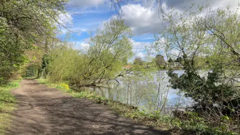 Poynton Pool looks like a small lake. A line of trees lies on its banks. with a dusty, dirt path running along one of its sides.