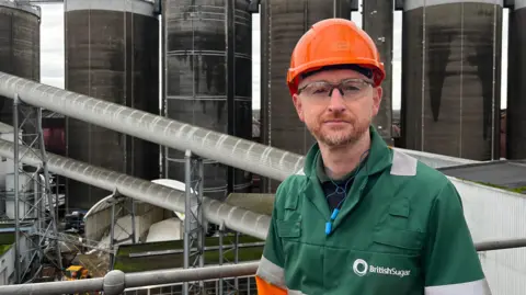 A man dressed in a green, orange and grey jacket along with safety glasses and an orange hard hat in front of multiple factory silos in a row