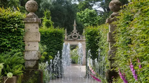 Burghley House Two columns provide an entrance to a new section of garden. Hedges and vertical jets of a water fountain are beyond. In the distance is an ornate stone entrance.